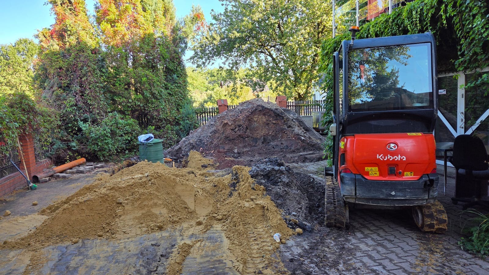 Mini koparka Kubota na posesji, obok hałdy ziemi i piasku. Widoczne ślady opon na kostce brukowej. W tle zieleń drzew i krzewów.