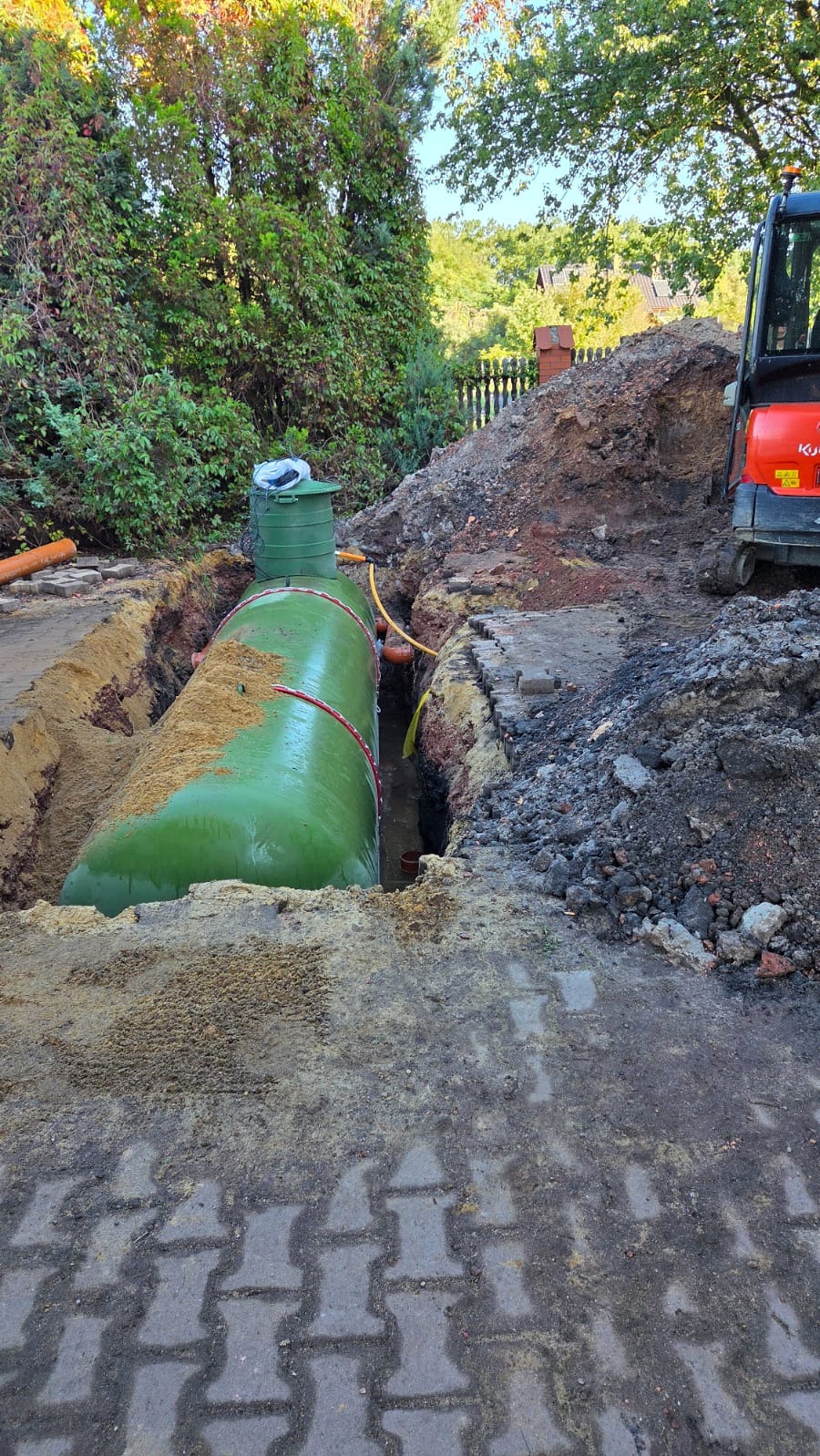 Zielony zbiornik na deszczówkę w wykopie, otoczony ziemią i kostką brukową. W tle widoczny mały traktor i zieleń. Instalacja zbiornika w trakcie prac.