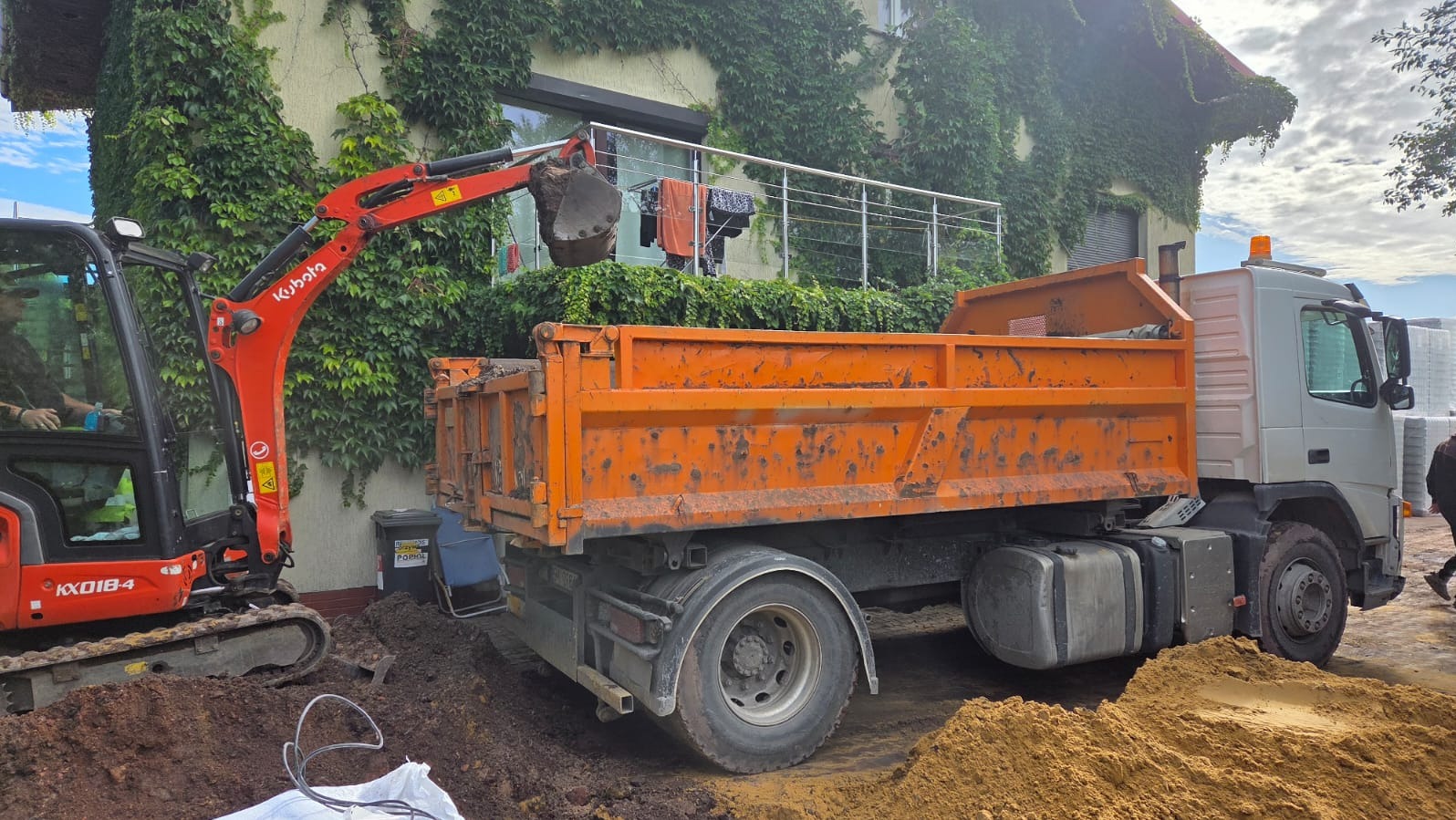 Pomarańczowa wywrotka i minikoparka Kubota przy domu z bluszczem podczas prac ziemnych. Łyżka koparki nad skrzynią ładunkową. Widoczny operator w kabinie.