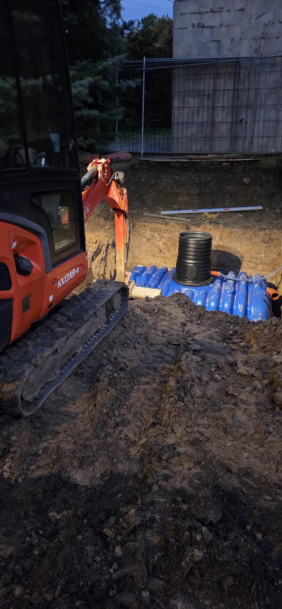 Mini koparka Kubota KX018-4 przy wykopie z niebieskimi zbiornikami retencyjnymi i czarną studzienką. Widoczne ślady opon w ziemi.