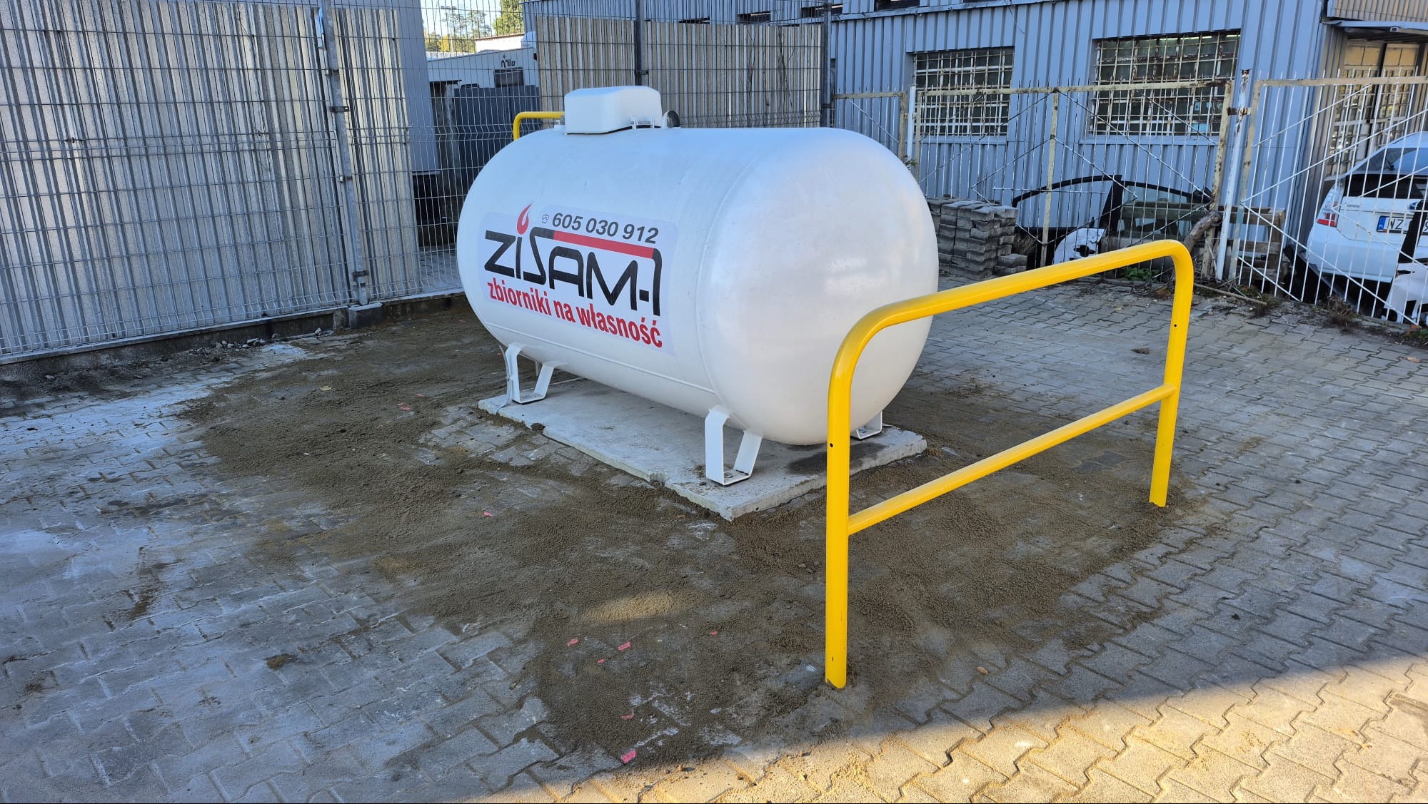 Biały zbiornik Zisam na betonowej podstawie, otoczony żółtą barierką. Teren wokół wyrównany piaskiem, w tle budynek z blachy falistej i metalowe ogrodzenie.