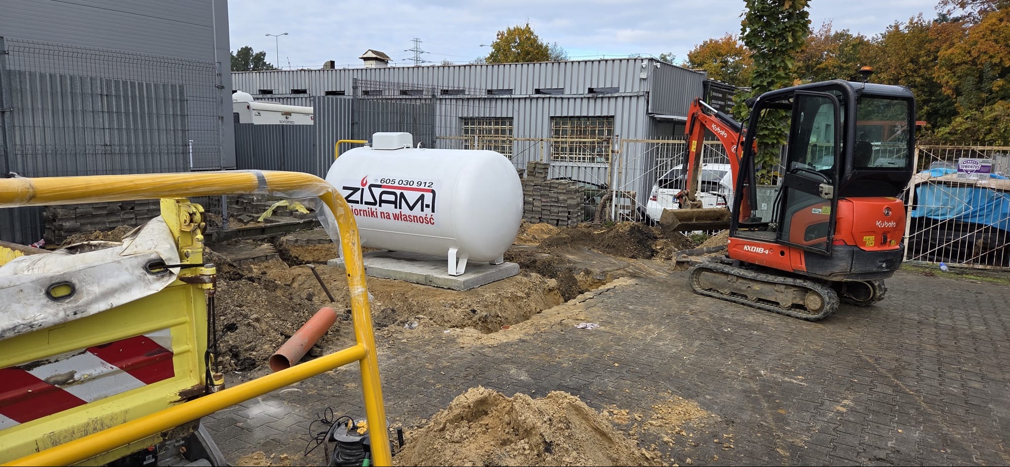 Montaż zbiornika gazowego ZISAM na posesji. Wykop pod instalację, mini koparka Kubota KX018-4 w tle. Widoczne elementy instalacji i kostka brukowa.