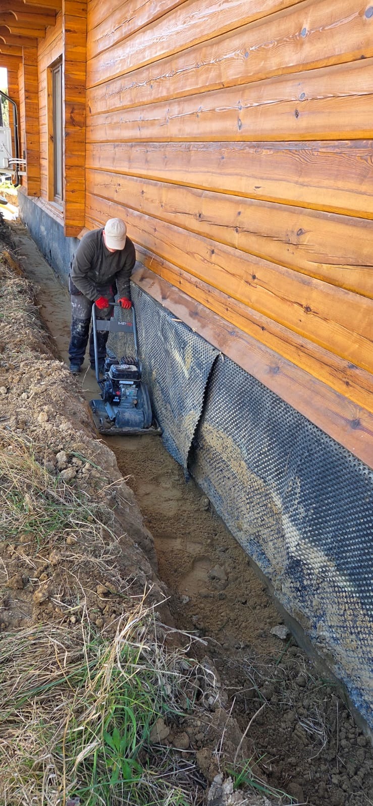 Pracownik zagęszcza grunt w wąskim wykopie wzdłuż drewnianego domu, zabezpieczonego folią kubełkową. Widoczne narzędzie pracy i fragment trawnika.