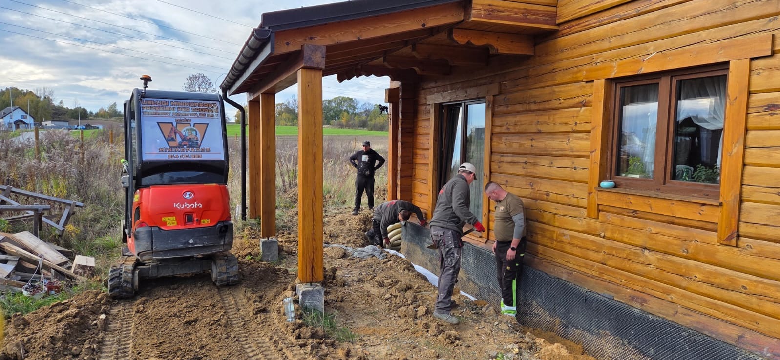 Prace ziemne przy drewnianym domu: układanie drenażu z użyciem minikoparki Kubota. Widoczni pracownicy i przygotowany wykop.