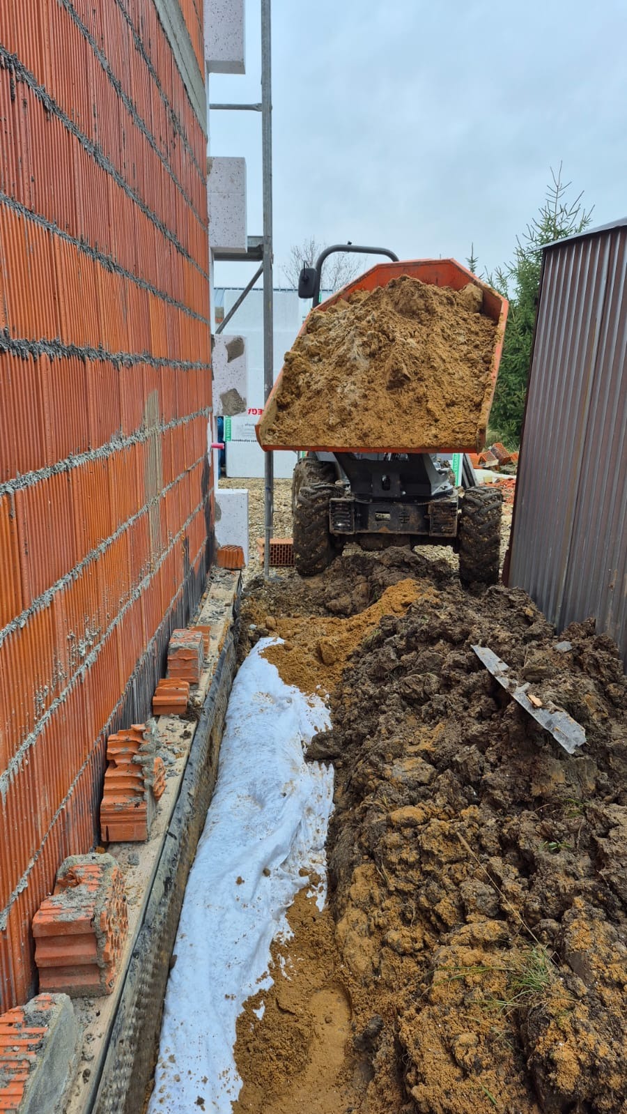 Wywóz ziemi w Wyry mini wywrotką obok fundamentów budynku z cegły. Wykop zabezpieczony geowłókniną. Prace ziemne w toku, widoczna pryzma ziemi.