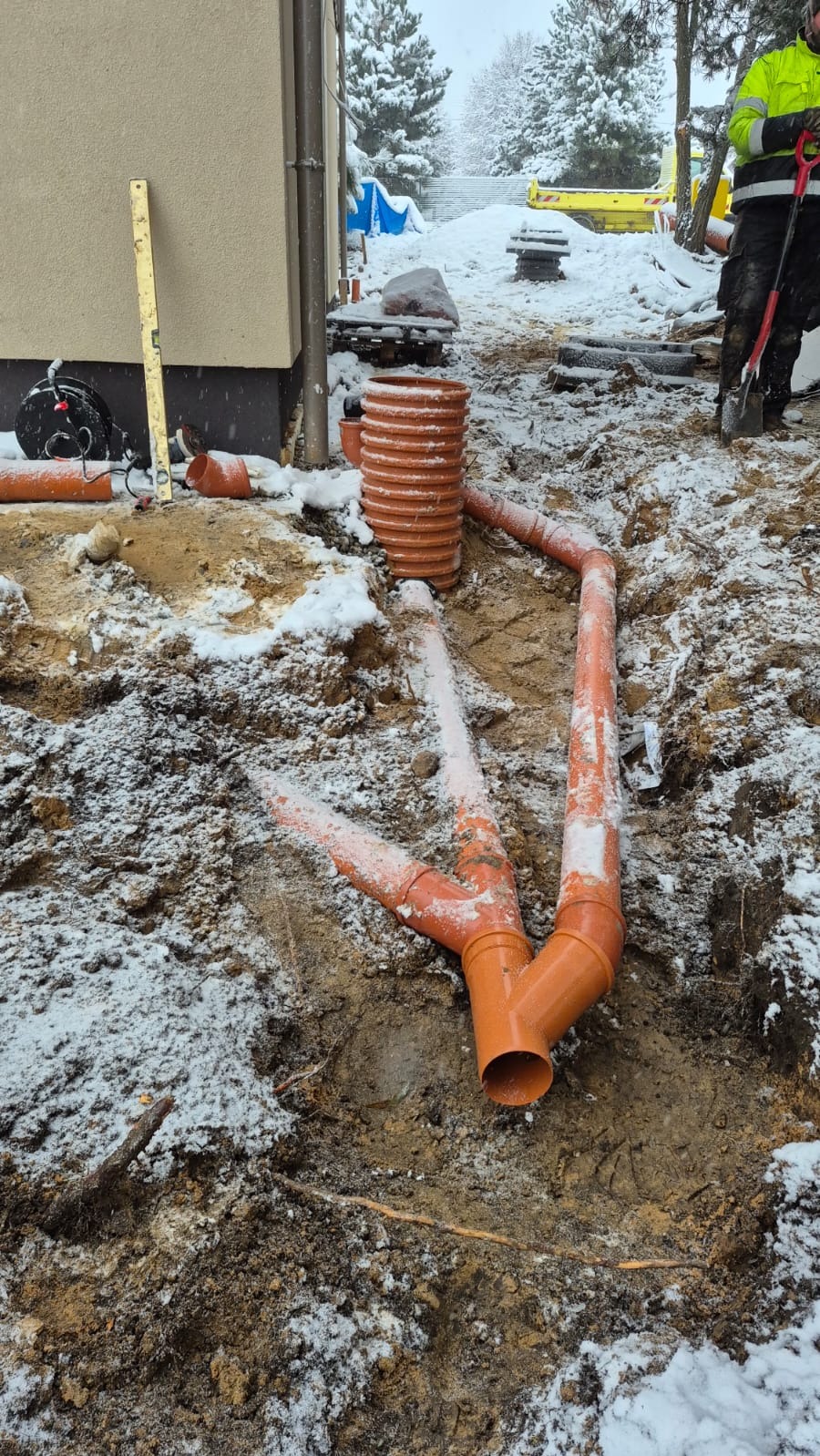 Montaż pomarańczowych rur drenażowych w wykopie pokrytym śniegiem, widoczny pracownik z łopatą i budynek w tle. Zimowa sceneria.