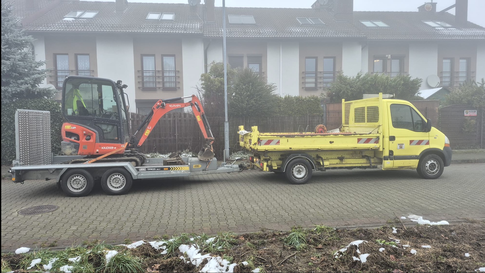 Żółta ciężarówka z przyczepą, na której stoi pomarańczowa mini koparka Kubota, zaparkowana na kostce brukowej w pochmurny dzień. W tle domy.