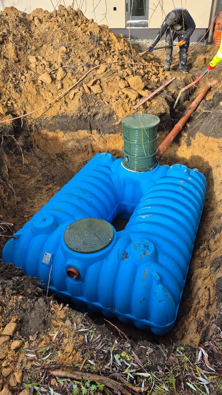 Niebieski zbiornik na szambo w wykopie, widoczne rury i pracownicy w tle. Ujęcie z góry, ziemia wokół zbiornika.