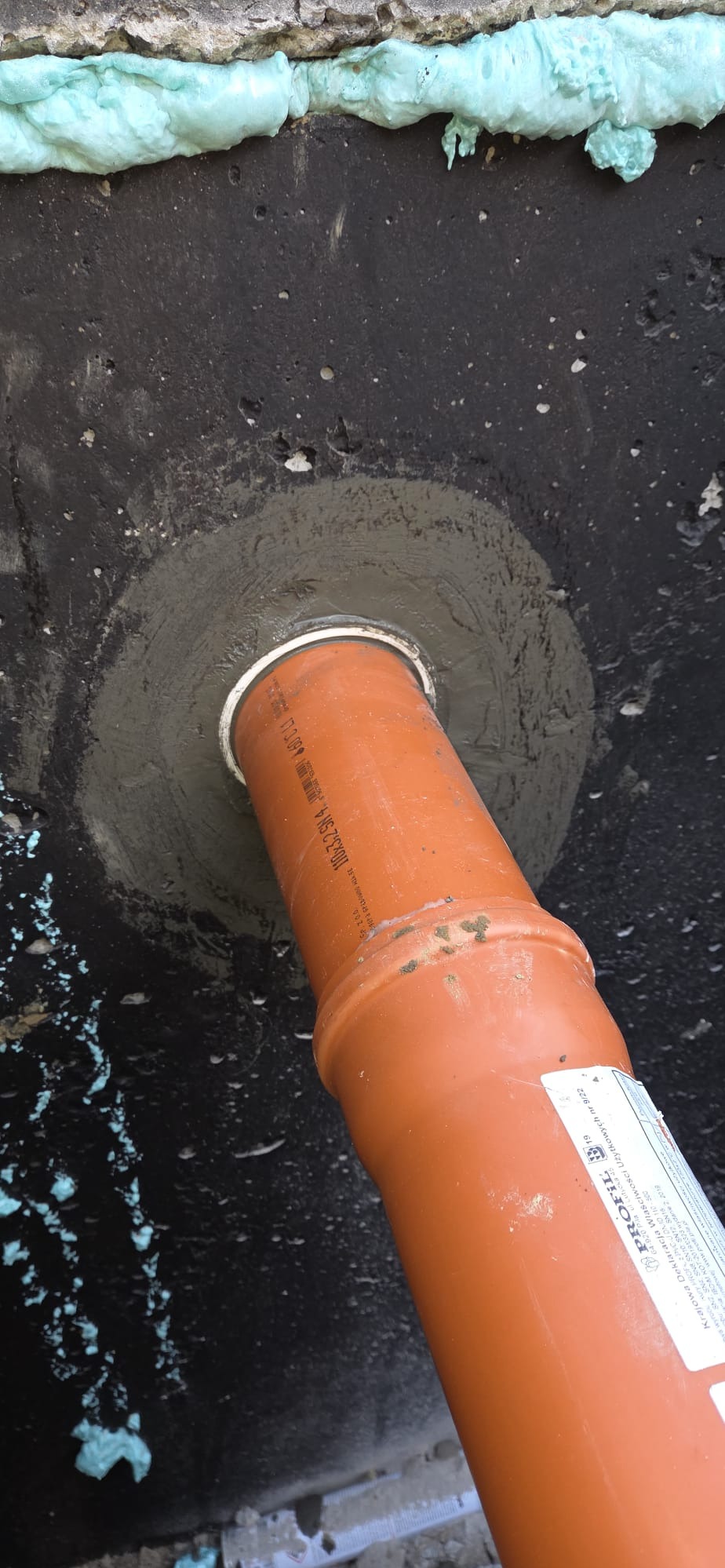 Pomarańczowa rura kanalizacyjna w otworze w czarnej, zabezpieczonej ścianie, uszczelniona szarym cementem. Widoczna pianka montażowa i etykieta na rurze.