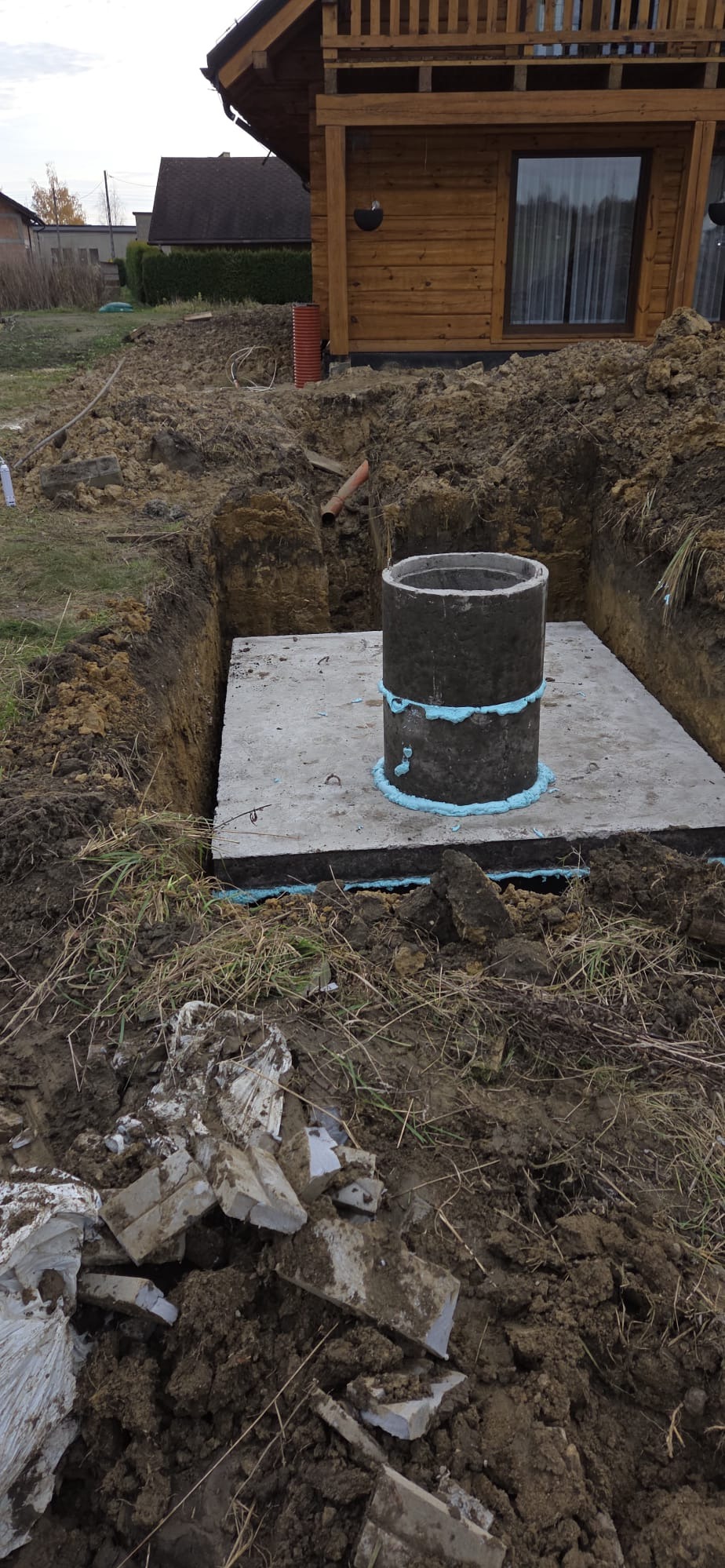 Betonowa studzienka kanalizacyjna w wykopie, zabezpieczona pianką montażową, na tle drewnianego domu i ziemi. Widoczne gruz i resztki materiałów budowlanych.