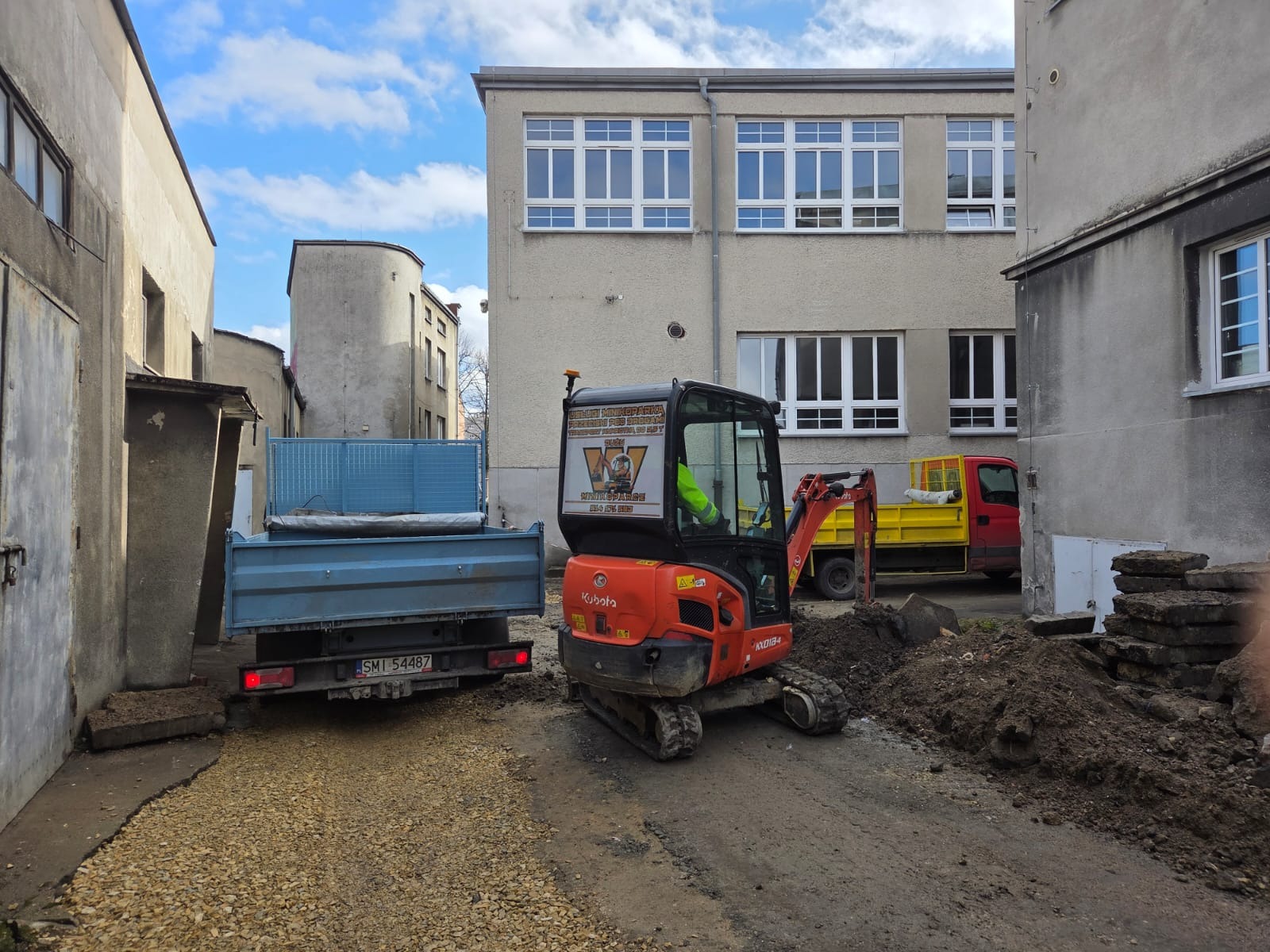 Pomarańczowa mini koparka Kubota w trakcie prac ziemnych na terenie budowy, obok zaparkowana ciężarówka z wywrotką. Widoczne budynki w tle.