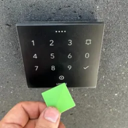 Czarny panel dotykowy z klawiaturą numeryczną i zielonym breloczkiem zbliżeniowym trzymanym w dłoni na tle szarej ściany.