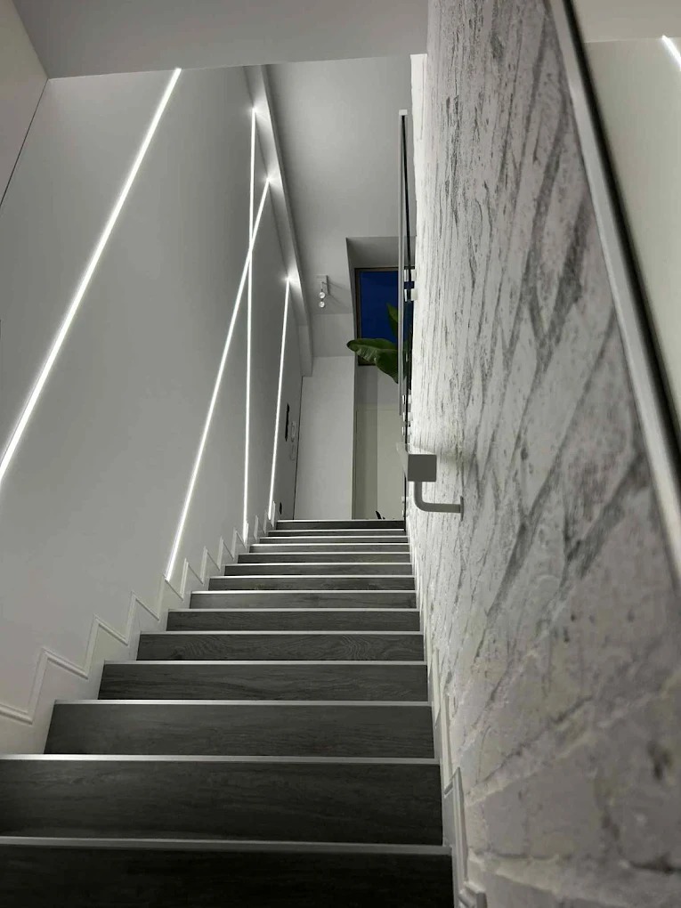 Nowoczesne schody z podświetleniem LED wzdłuż ściany i dekoracyjną cegłą. Minimalistyczny design z rośliną w tle.