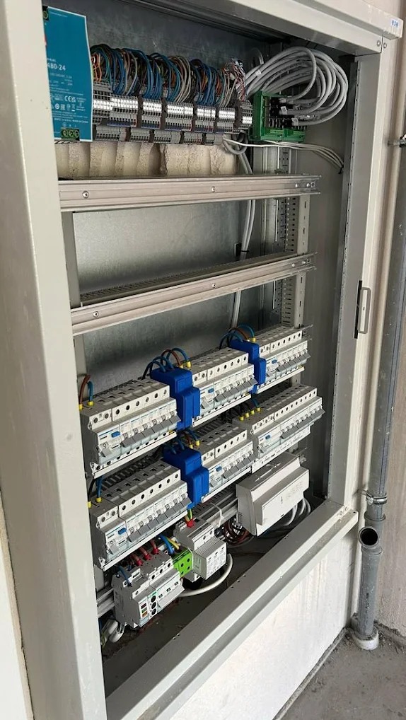 Rozdzielnica elektryczna z zainstalowanymi bezpiecznikami i okablowaniem w metalowej obudowie, widoczna pionowa rura instalacyjna po prawej stronie.