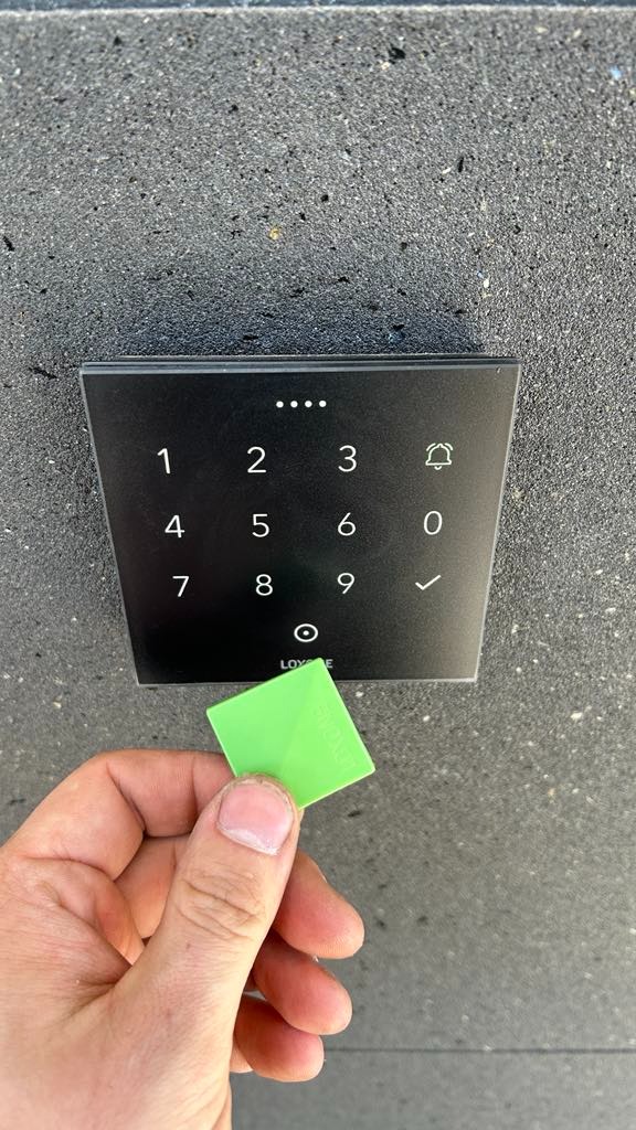 Czarny panel dotykowy z klawiaturą numeryczną i zielonym breloczkiem zbliżeniowym trzymanym w dłoni na tle szarej ściany.