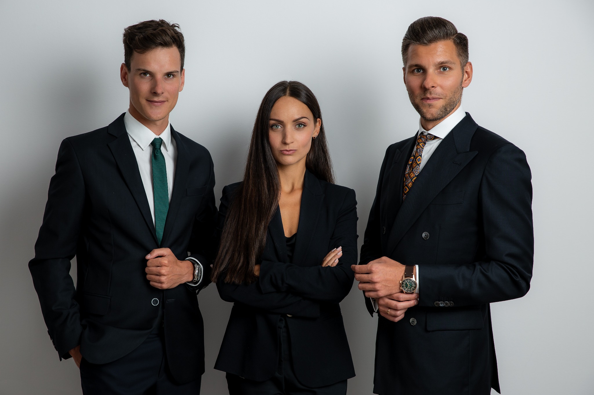 Profesjonalne portretowe ujęcie trzech osób w garniturach na jasnym tle. Kobieta pośrodku ze skrzyżowanymi rękami. Mężczyźni po bokach z nonszalancką elegancją.
