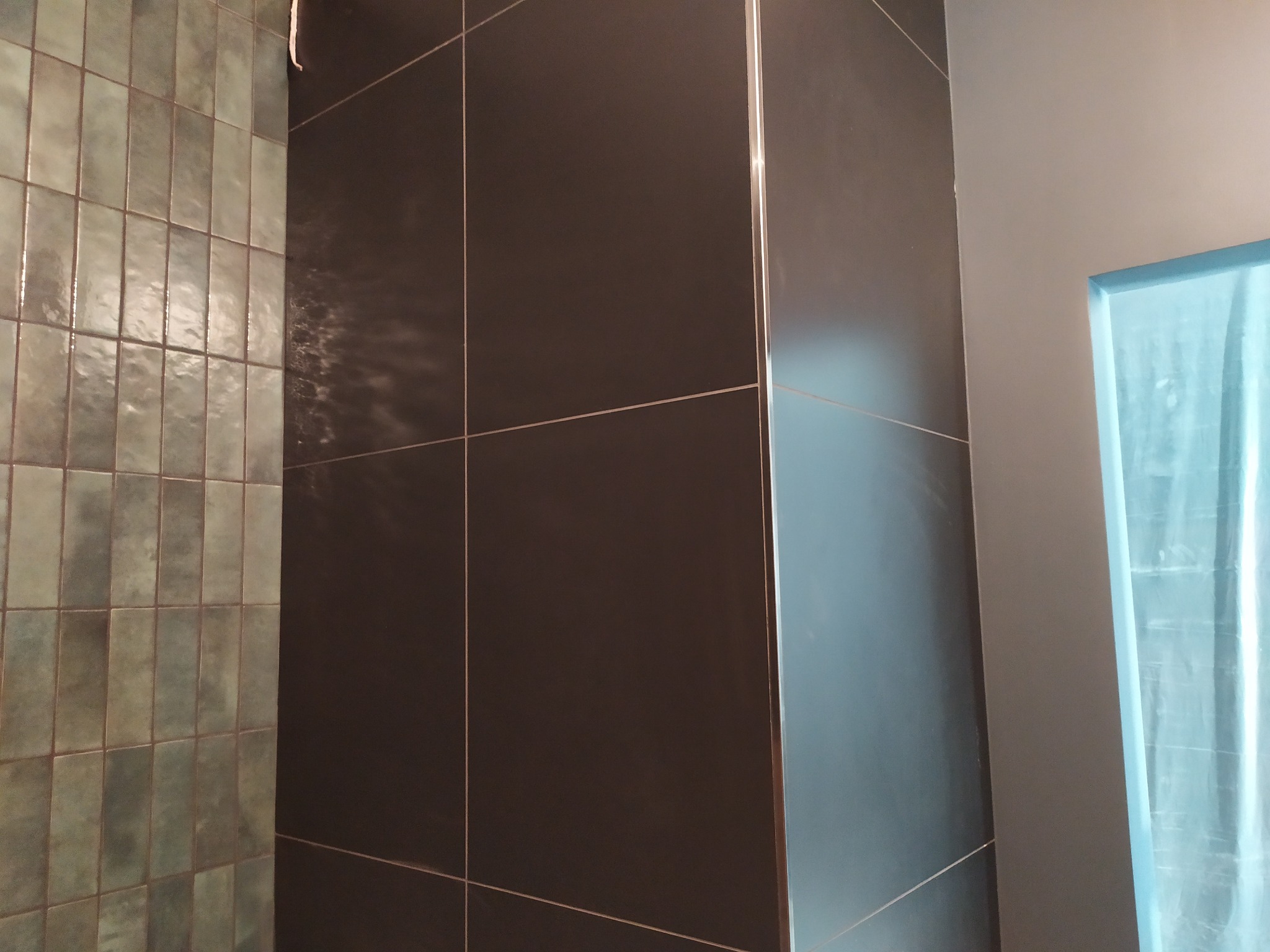 Wykończona ściana w łazience z ciemnymi, kwadratowymi płytkami i fragmentem ściany z wąskimi, zielonkawymi płytkami. Widoczny fragment zabezpieczonego folią otworu.