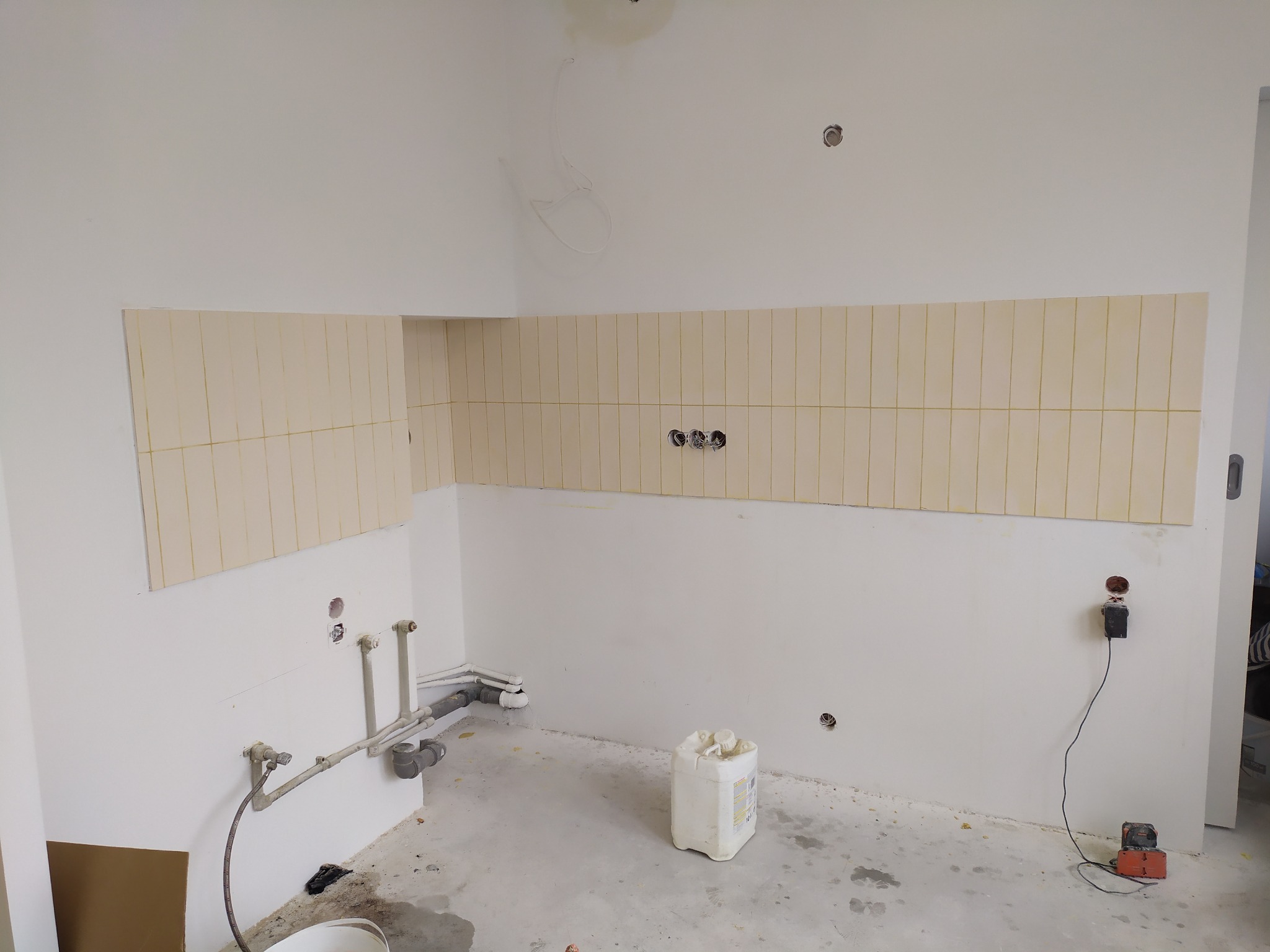 Fragment kuchni w trakcie prac wykończeniowych: częściowo położone beżowe płytki na ścianie, widoczne instalacje i narzędzia. W tle białe ściany.