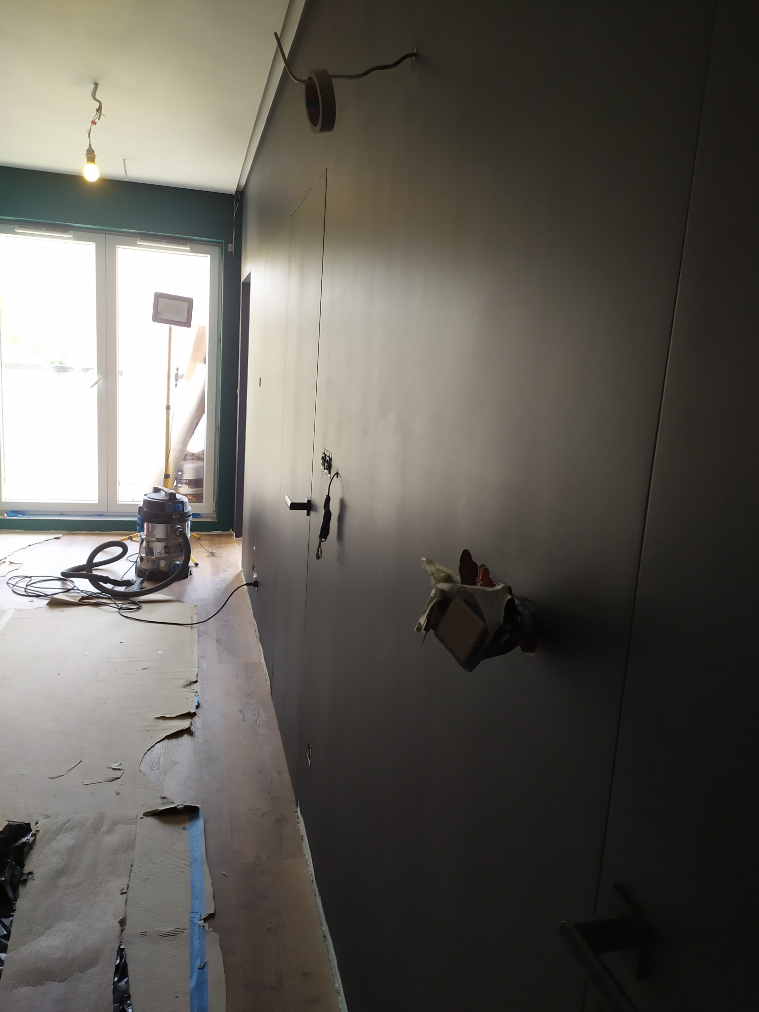 Wnętrze w trakcie remontu: ciemnoszara ściana z puszką elektryczną, podłoga zabezpieczona kartonem, odkurzacz przemysłowy w tle. Widoczne niedokończone prace.