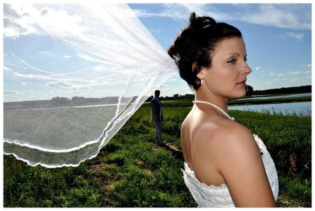 Panna młoda w białej sukni i perłach, profil, welon powiewający na wietrze, w tle mężczyzna w garniturze i krajobraz z wodą i zielenią, plener.