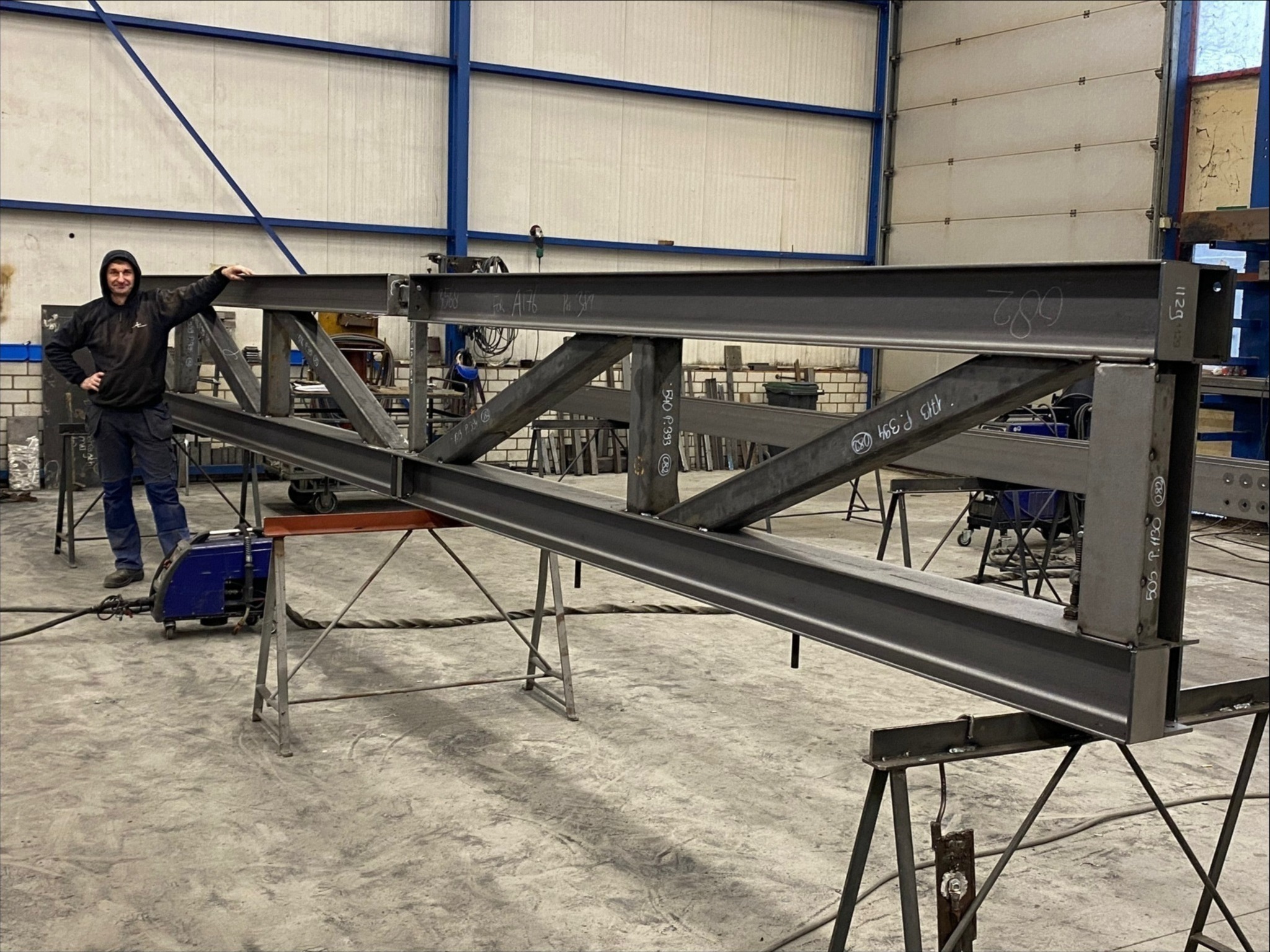 Konstrukcja metalowa spawana w warsztacie, prawdopodobnie element bramy lub ogrodzenia, z pracownikiem opierającym się o nią, obok spawarka.