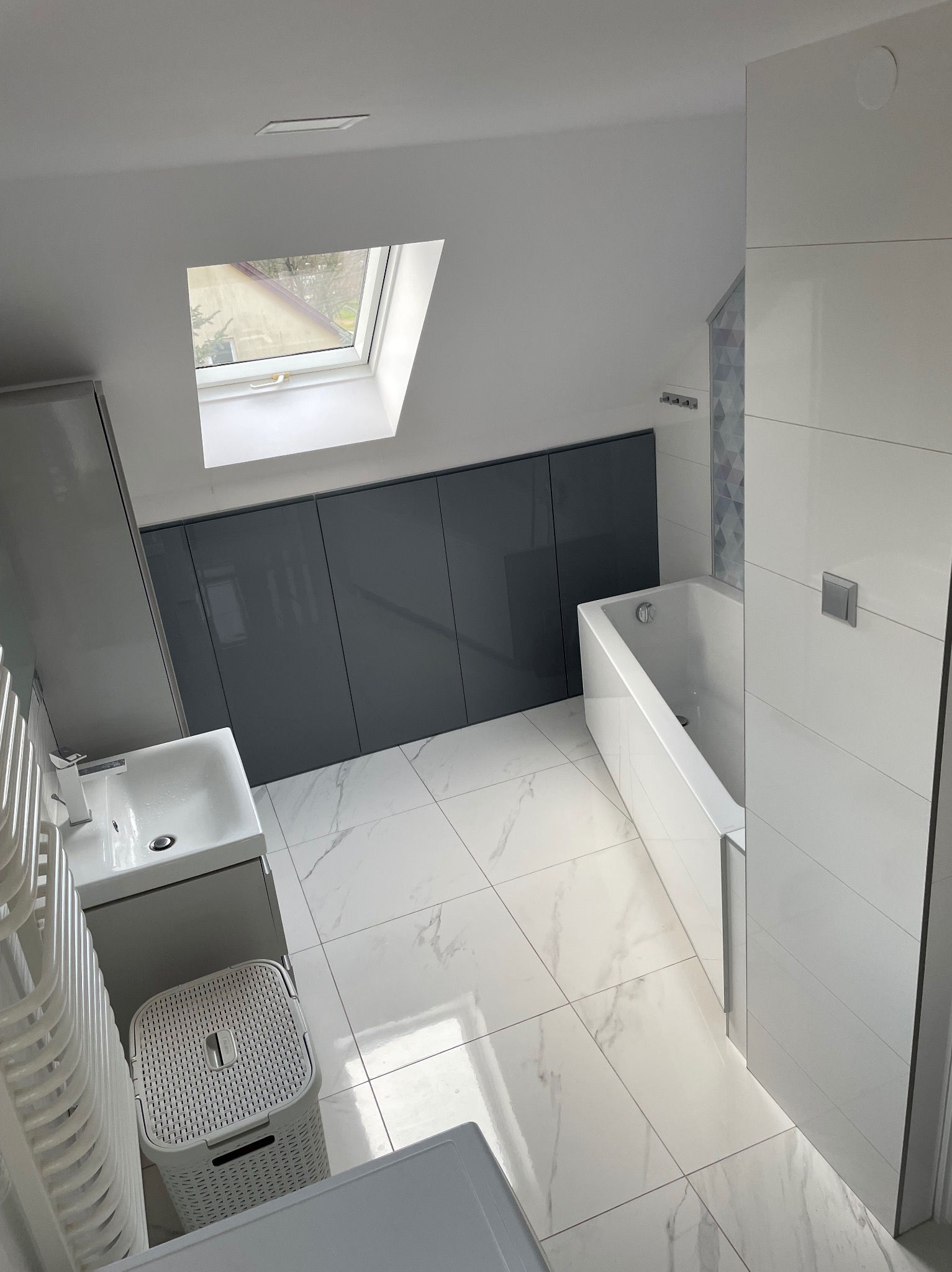 Nowoczesna łazienka na poddaszu z oknem dachowym, biało-szara kolorystyka, marmurowe płytki podłogowe, zabudowa szafek pod skosem, minimalistyczna armatura.