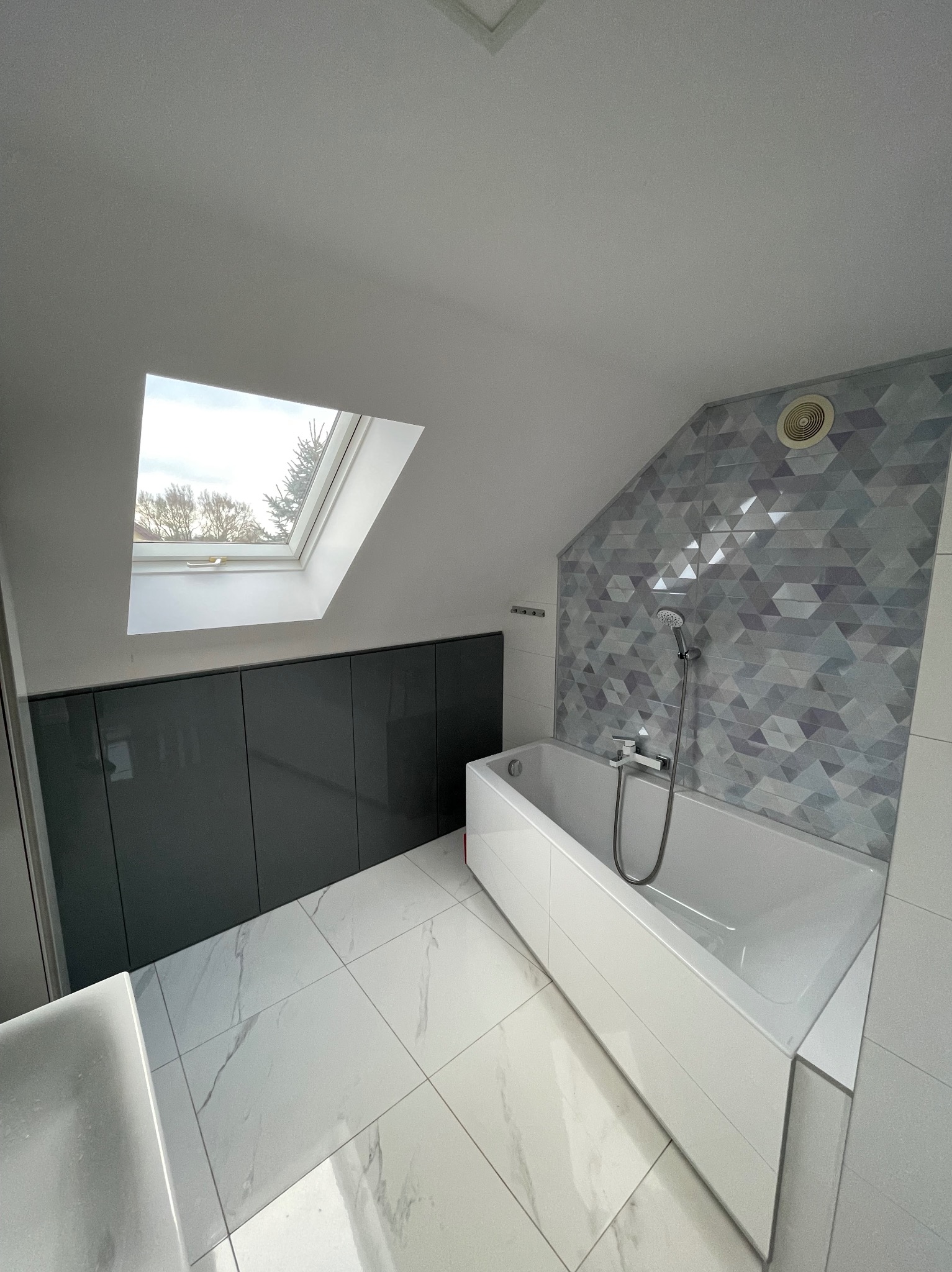 Nowoczesna łazienka na poddaszu z białą wanną, szarą mozaiką na ścianie i błyszczącymi, marmurkowymi płytkami podłogowymi. Okno dachowe wpuszcza naturalne światło.