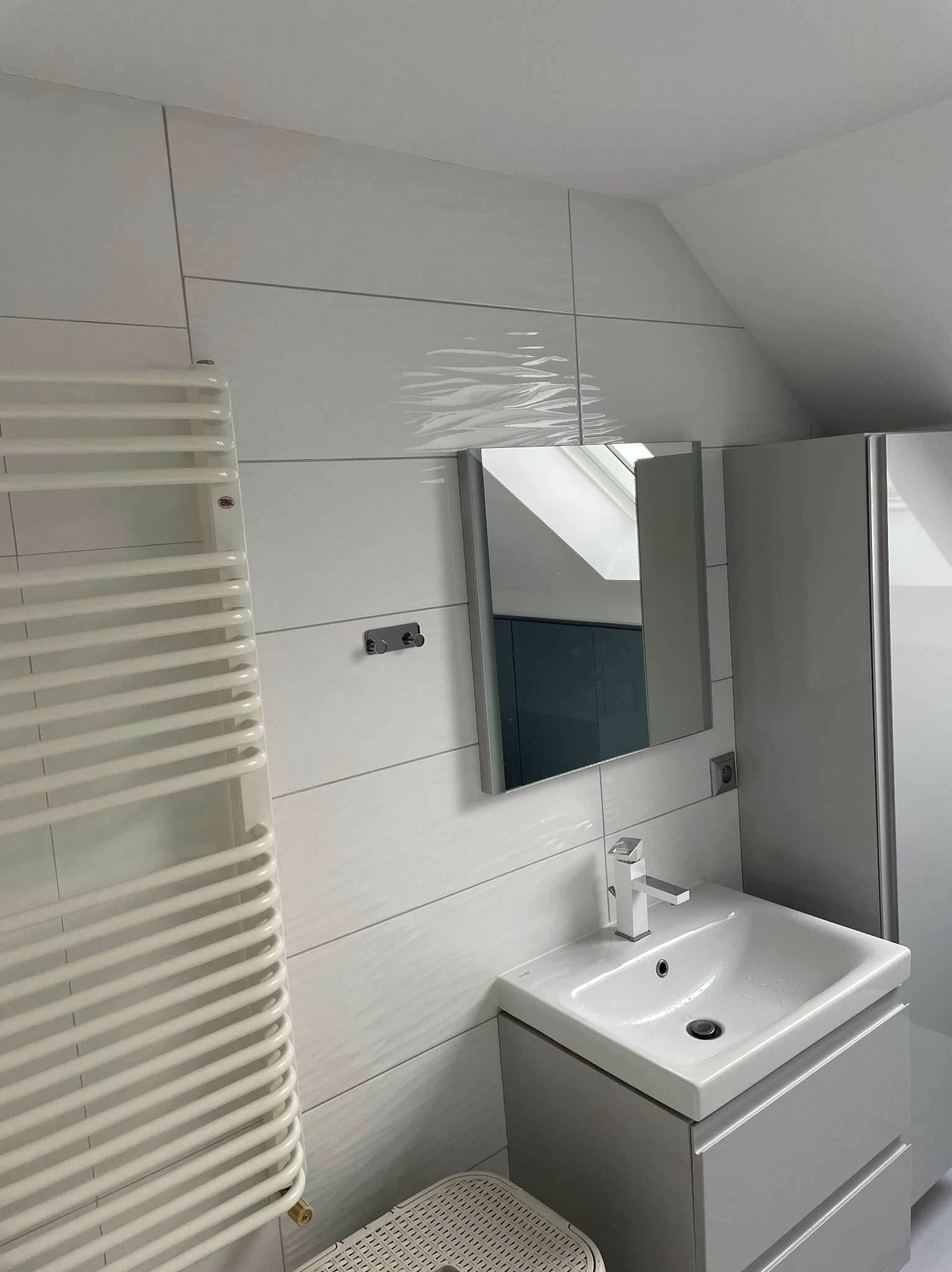 Biała łazienka na poddaszu z grzejnikiem drabinkowym, lustrem i umywalką z szafką. Płytki imitujące fale. Widoczny fragment kabiny prysznicowej.