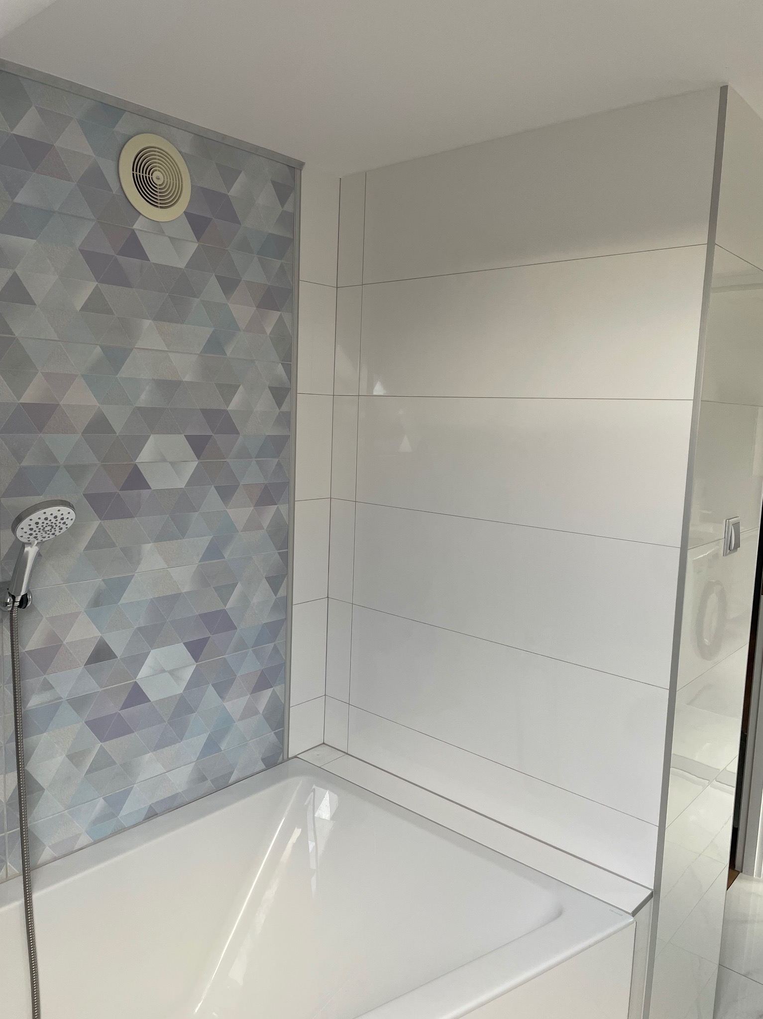 Wykończona łazienka z białą wanną, geometrycznymi płytkami w odcieniach szarości i białymi, poziomymi płytkami. Widoczny wentylator i słuchawka prysznicowa.