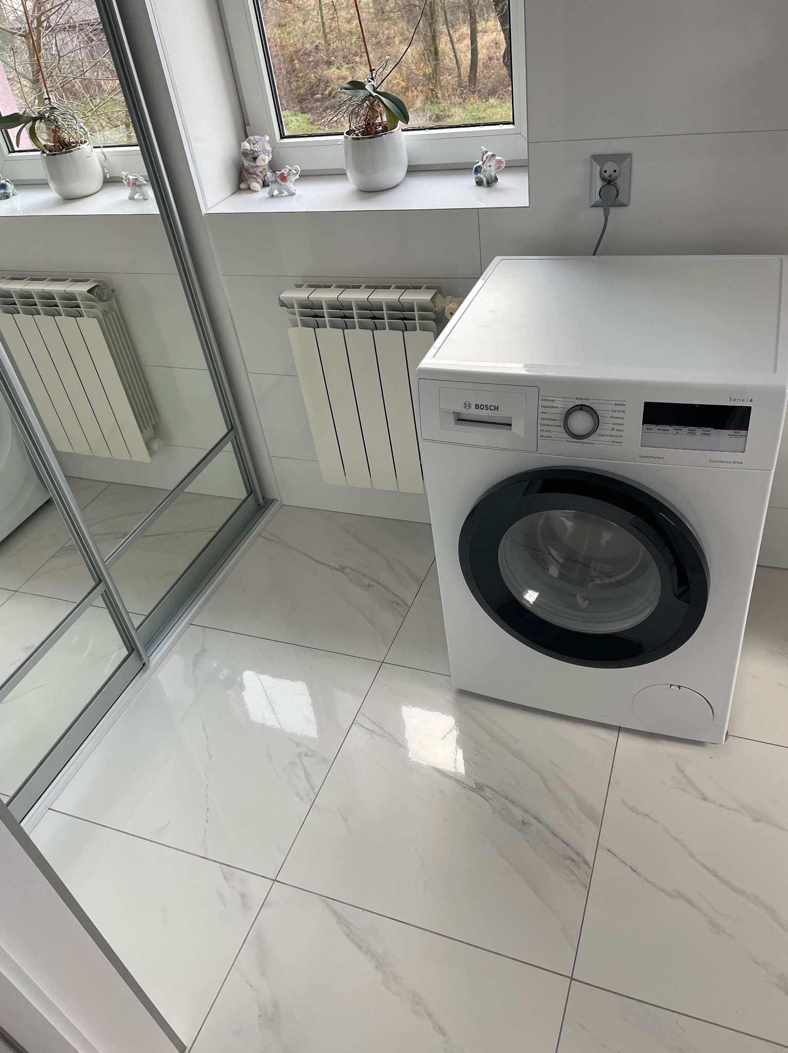 Biała łazienka z marmurową podłogą i pralką Bosch Serie 4. Widok na okno z dekoracjami i grzejnik. Nowoczesne wykończenie, jasne wnętrze.