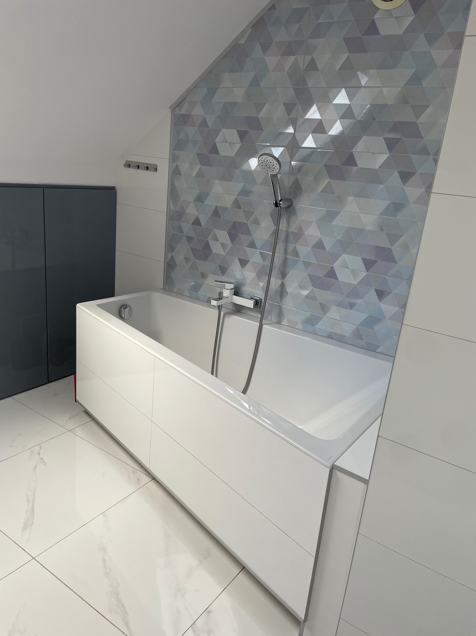 Nowoczesna łazienka na poddaszu: biała wanna obudowana płytkami, geometryczna mozaika na ścianie, marmurowe płytki podłogowe i szara szafka w tle.