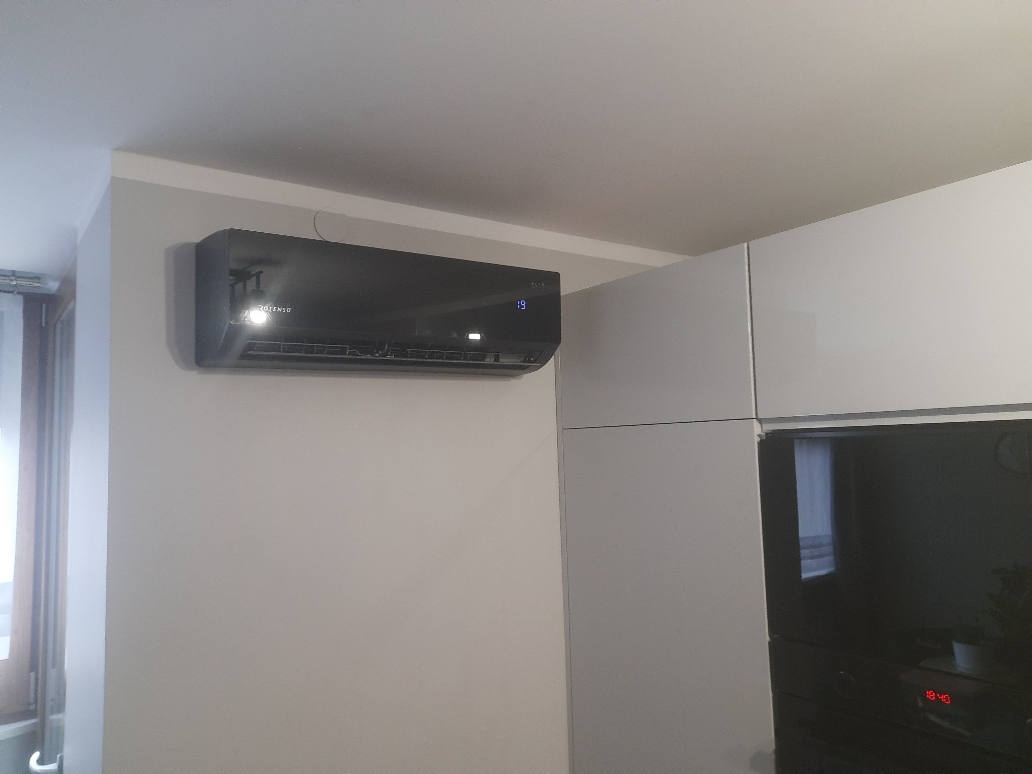 Czarna jednostka wewnętrzna klimatyzacji Rotenso, zamontowana na ścianie w nowoczesnym wnętrzu, z widocznym wyświetlaczem temperatury. Obok zabudowa kuchenna.