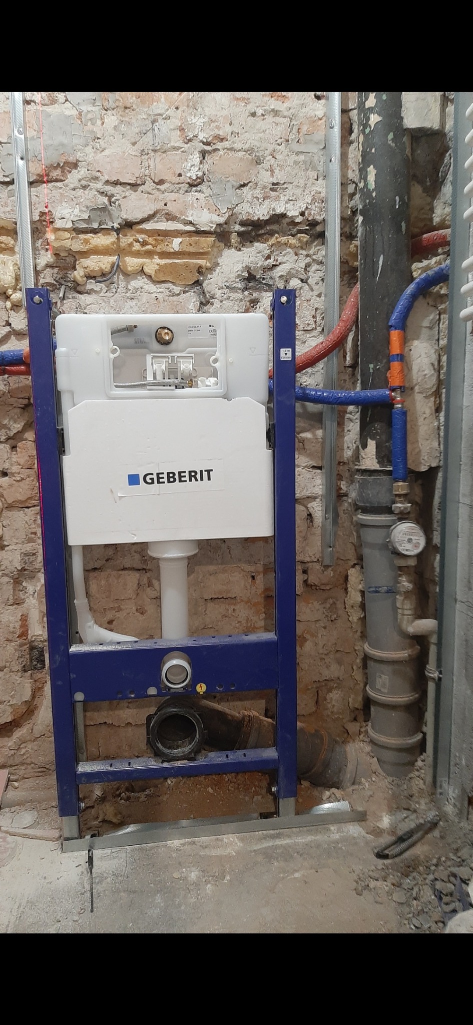 Stelaż podtynkowy Geberit zamontowany na tle starej, ceglanej ściany w trakcie remontu łazienki. Widoczne rury i elementy instalacji wodno-kanalizacyjnej.