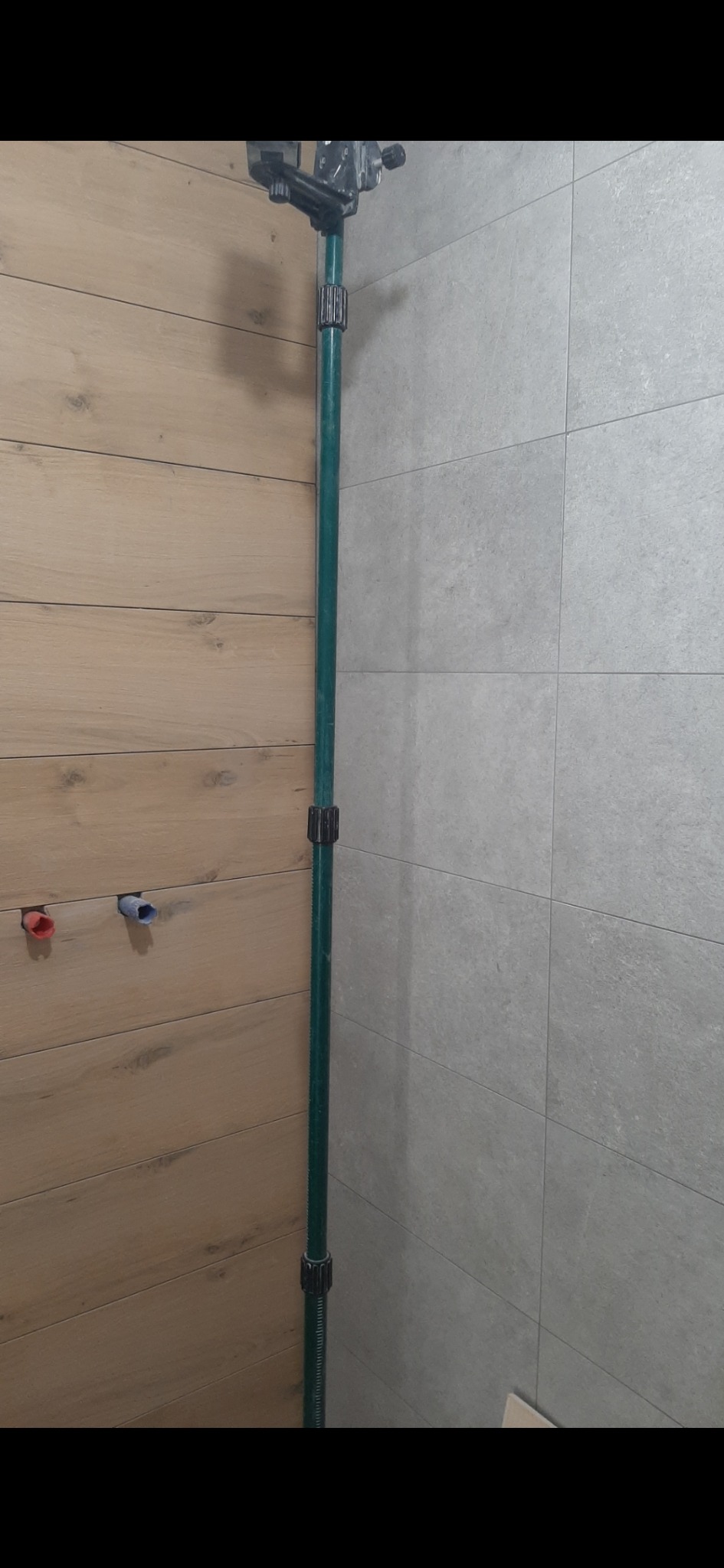 Wykończona łazienka z drewnopodobnymi i szarymi płytkami. Widoczny pionowy niwelator laserowy i wystające rury instalacji wodnej w ścianie.