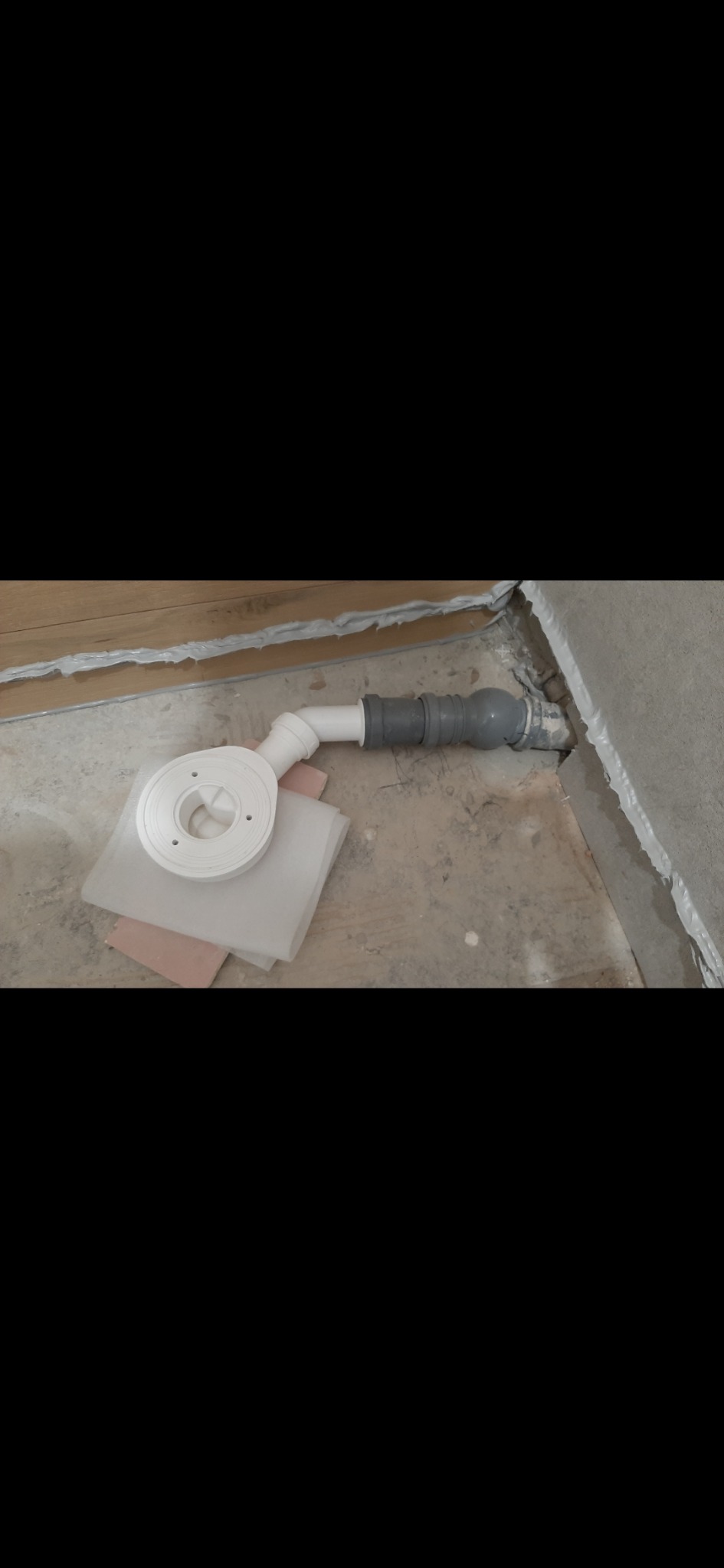 Odpływ liniowy z rurami w surowej łazience, widoczne materiały izolacyjne. Detal instalacji przed wykończeniem podłogi. Ujęcie z bliska, szare płytki w tle.