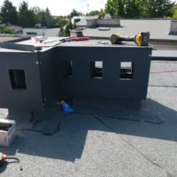 Obróbka blacharska na dachu z papy, narzędzia dekarskie i krem z filtrem UV. Blacha w kolorze grafitowym, przymocowana na nity. W tle zieleń drzew.