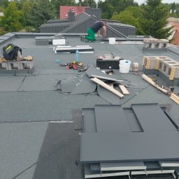 Jarosław Farfał - Dekarz w trakcie pracy na płaskim dachu, układa szare płyty. Widoczne narzędzia, materiały budowlane i fragmenty starego pokrycia. W tle drzewa i zabudowania.