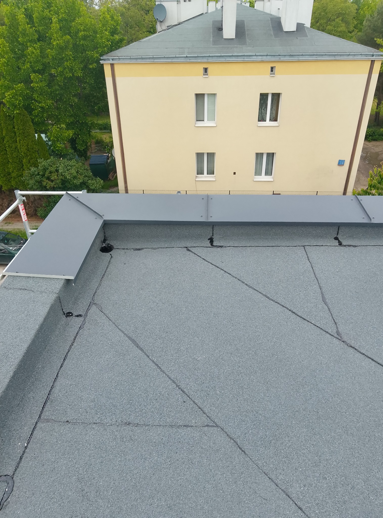 Szary dach pokryty papą termozgrzewalną z obróbkami blacharskimi w kolorze szarym. W tle budynek mieszkalny i drzewa.