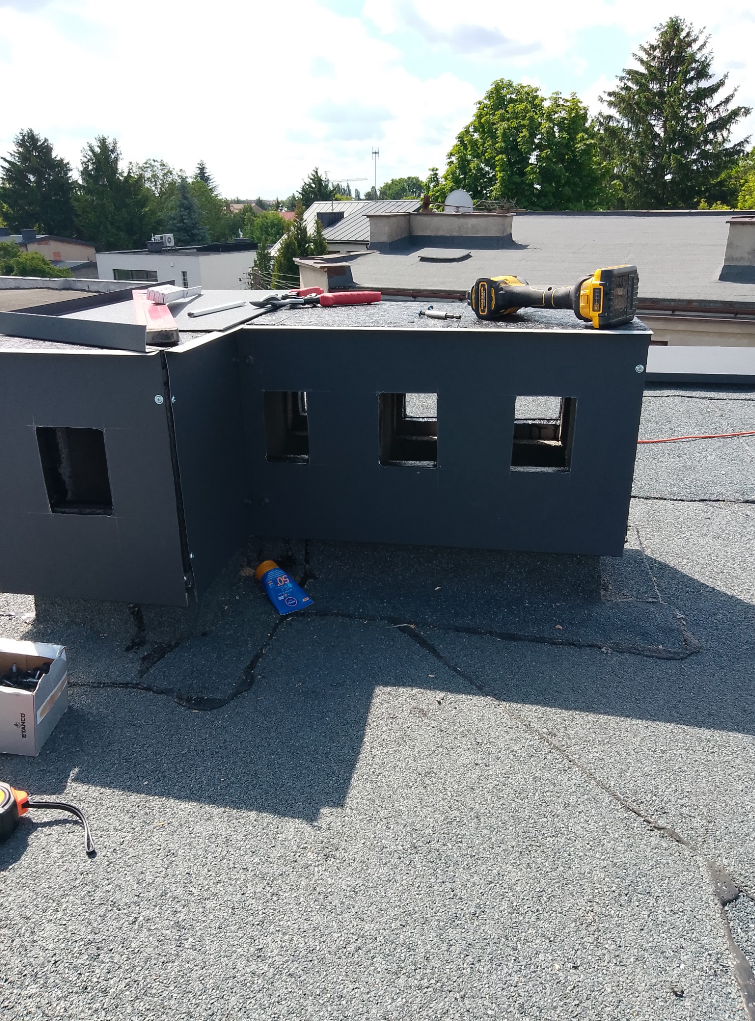 Obróbka blacharska na dachu z papy, narzędzia dekarskie i krem z filtrem UV. Blacha w kolorze grafitowym, przymocowana na nity. W tle zieleń drzew.