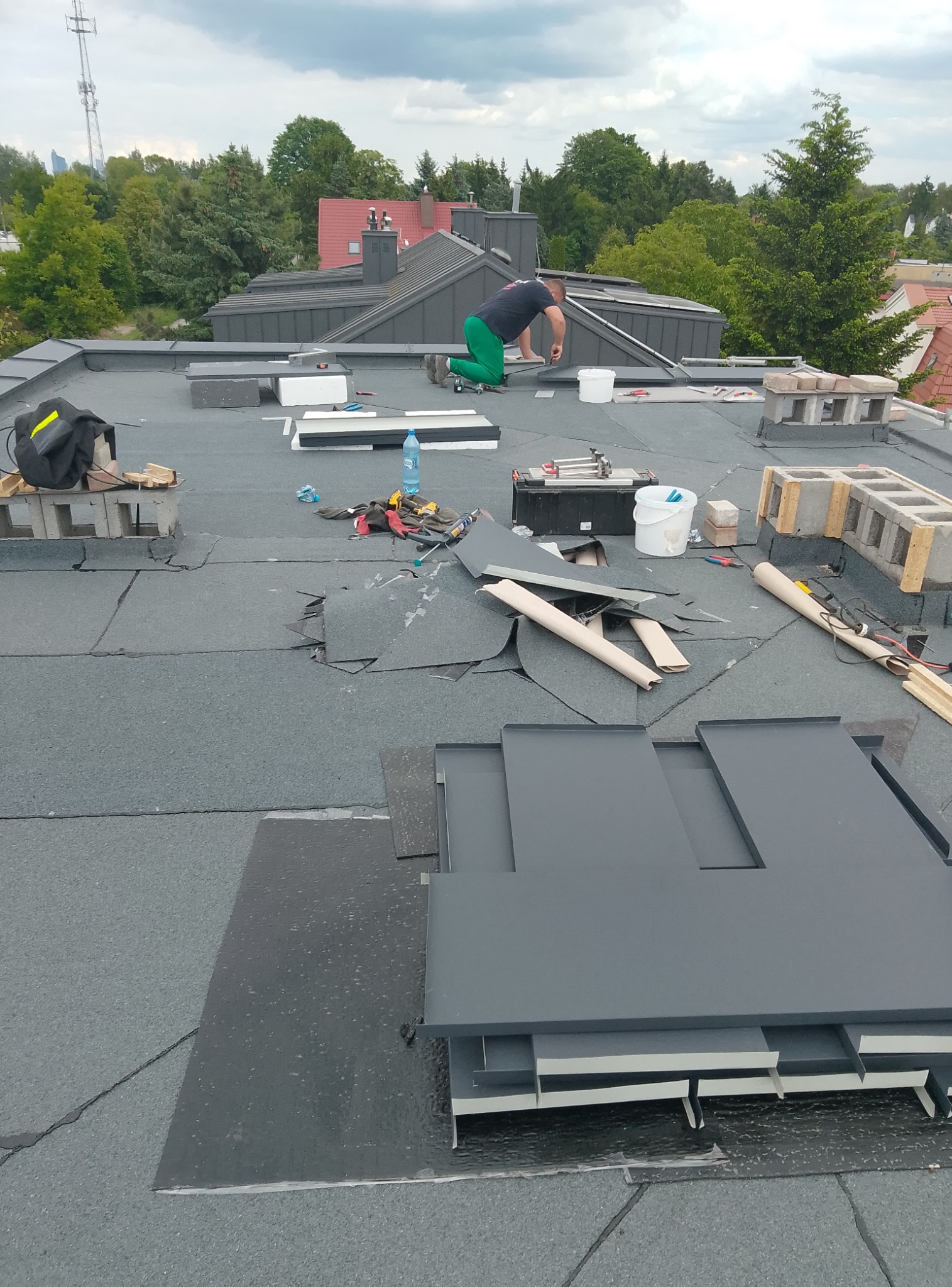 Dekarz w trakcie pracy na płaskim dachu, układa szare płyty. Widoczne narzędzia, materiały budowlane i fragmenty starego pokrycia. W tle drzewa i zabudowania.