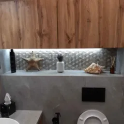 Nowoczesna łazienka z drewnianymi szafkami, szarą mozaiką i dekoracjami: muszla, rozgwiazda, wazon z lawendą. Oświetlenie LED podkreśla geometryczny wzór płytek.