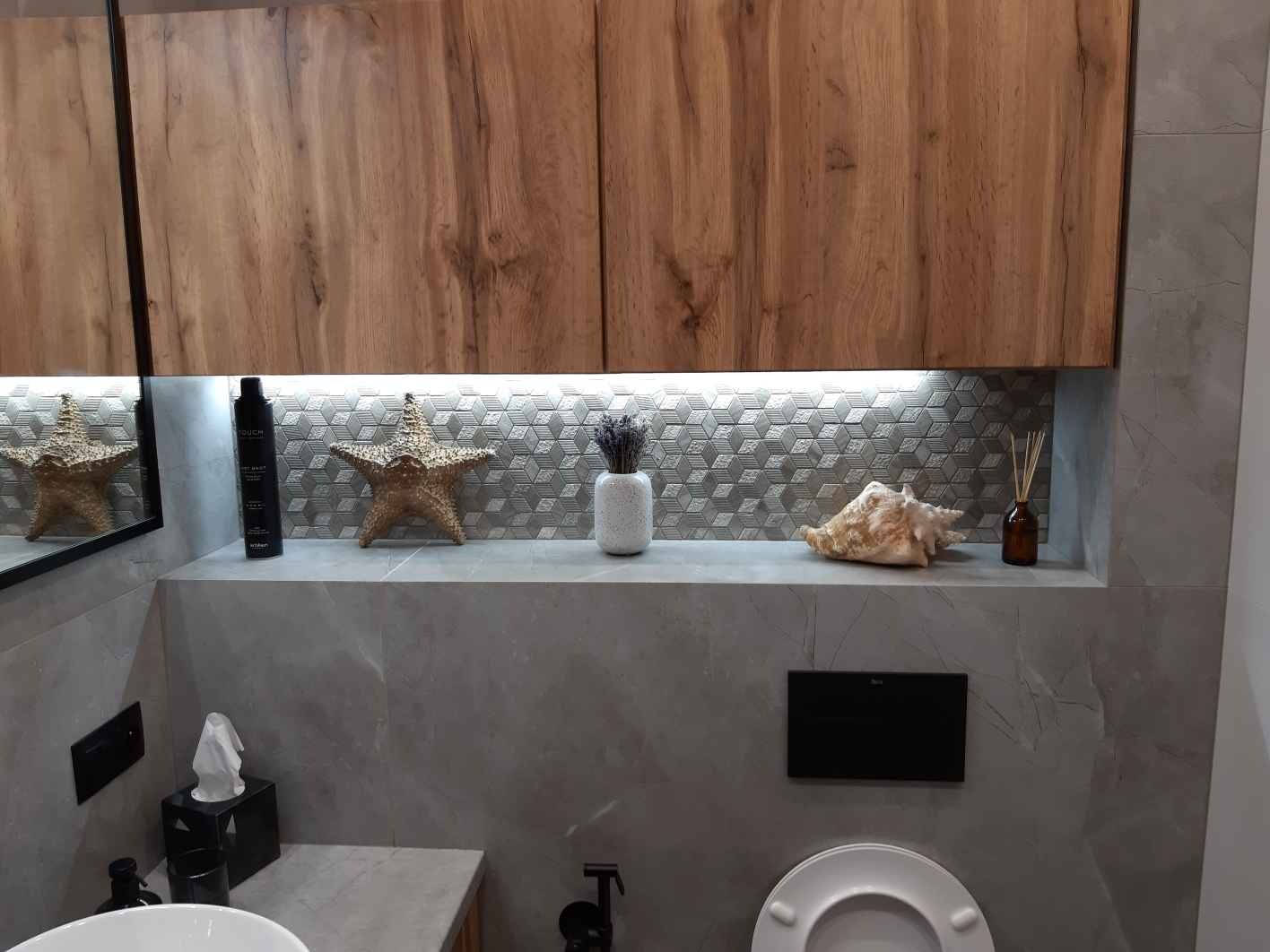 Nowoczesna łazienka z drewnianymi szafkami, szarą mozaiką i dekoracjami: muszla, rozgwiazda, wazon z lawendą. Oświetlenie LED podkreśla geometryczny wzór płytek.