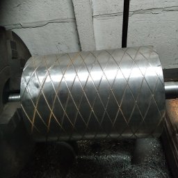 Skrawmet-plast - Metalowy wałek z diamentowym wzorem w tokarce, otoczony wiórami. Detal obróbki skrawaniem, widoczne ślady pracy.