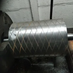 Metalowy wałek z diamentowym wzorem w tokarce, otoczony wiórami. Detal obróbki skrawaniem, widoczne ślady pracy.