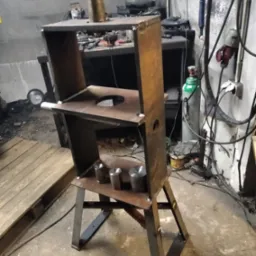 Metalowa konstrukcja spawana z profili, widoczne spawy, otwory i elementy cylindryczne. Surowy warsztat z butlą gazową w tle.