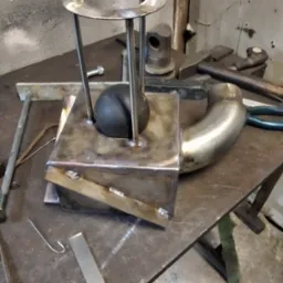 Spawany metalowy element z rurką, kulką i pierścieniem na warsztatowym stole. Widoczne ślady spawania i narzędzia.