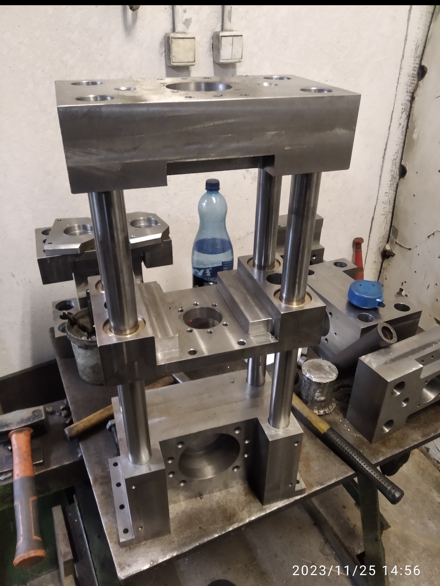 Stalowa konstrukcja z otworami i cylindrycznymi wspornikami na warsztatowym stole, obok narzędzia i butelka wody. Element obróbki metalu w surowym stanie.