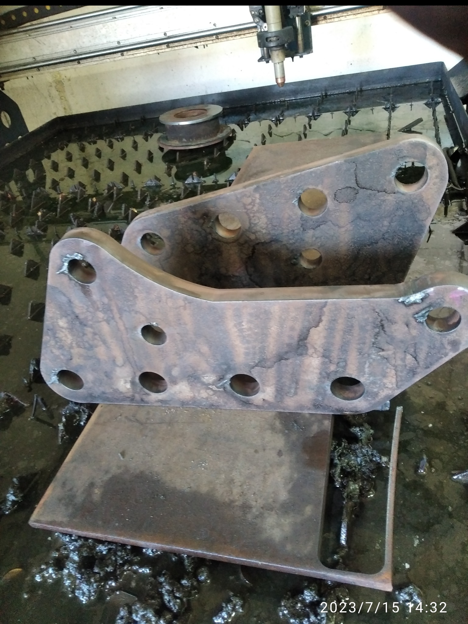 Dwa metalowe elementy z otworami, ułożone na metalowej płycie w maszynie do cięcia plazmowego. Widoczne ślady obróbki i chłodziwa.