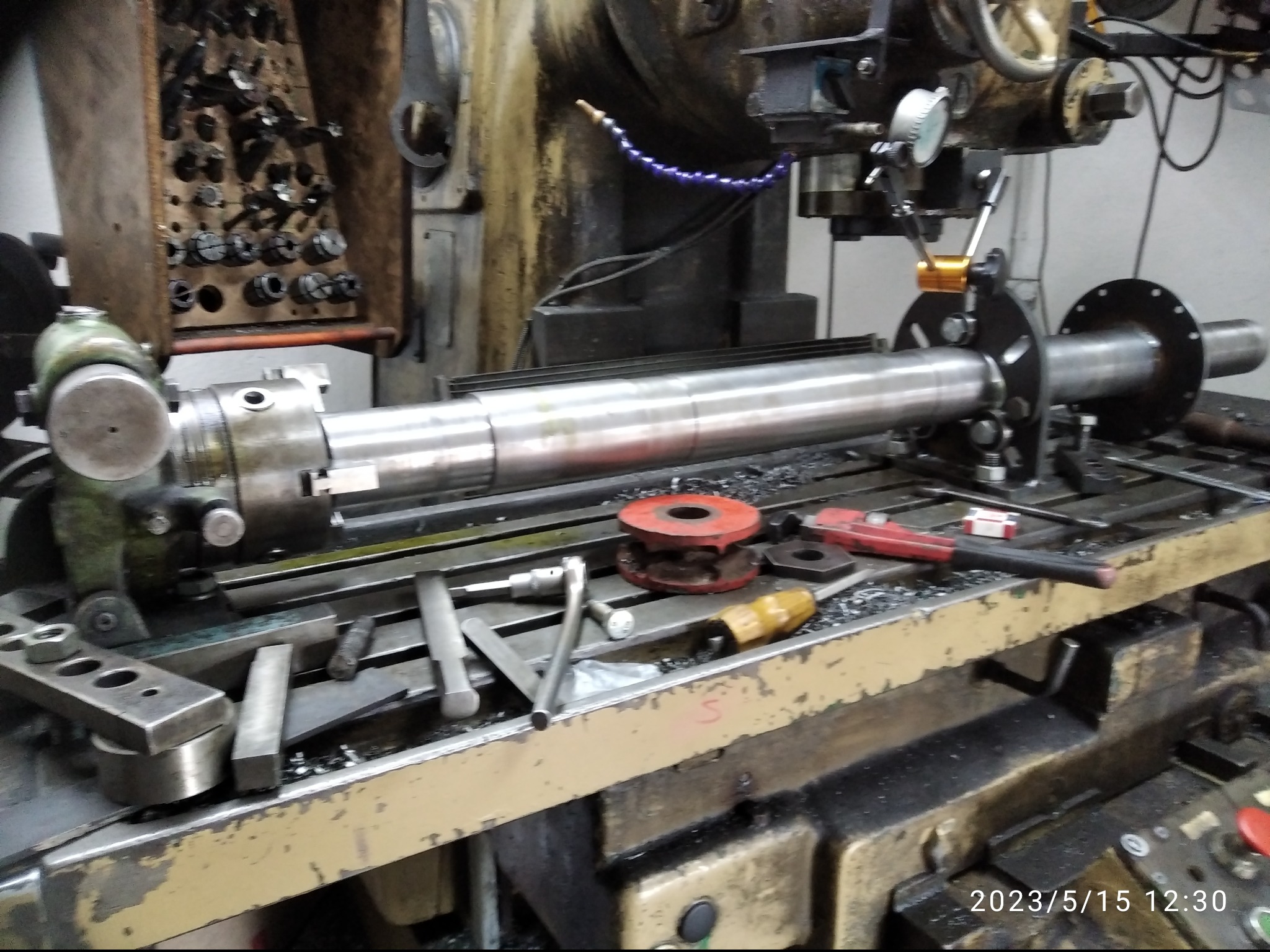 Metalowy wał zamocowany w tokarce, narzędzia i wióry na stole. Widoczne uchwyty tokarskie i wskaźnik zegarowy. Sprzęt do obróbki metalu w warsztacie.