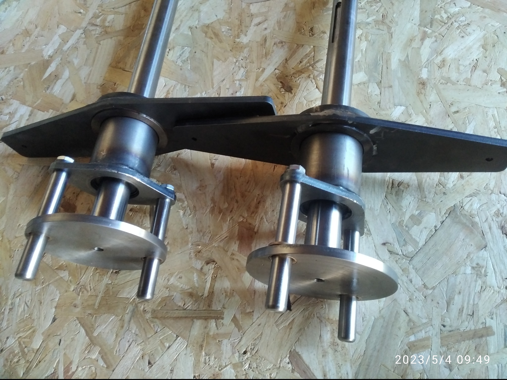 Dwa metalowe elementy z wałami i podstawami, prawdopodobnie wykonane w Kawęczynie, na tle płyty OSB. Widoczne ślady spawania.